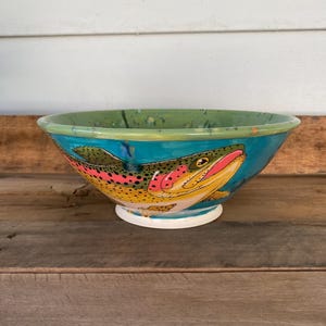 May include: A ceramic bowl with a hand-painted trout design. The bowl features a vibrant color palette with a turquoise background, a green rim, and a white base. The trout is depicted with yellow, pink, and black details.