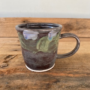 May include: A dark brown and green ceramic mug with a white rim. The mug has a unique glaze pattern with swirls of green and brown.