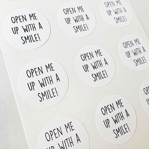 May include: A sheet of white round stickers with the text "Open me up with a smile!" printed in black.