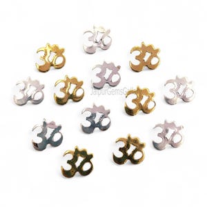 May include: A collection of small, metallic Om symbols in gold and silver tones. The symbols are scattered across a white surface, with varying levels of shine and detail. The image is well-lit, highlighting the intricate design of each charm.