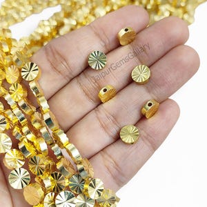 May include: A collection of small, round, gold-colored beads with a sunburst-like radial texture. Several individual beads are scattered, while others are strung together in long, vertical strands. The text JaipurGemsGallary is visible.