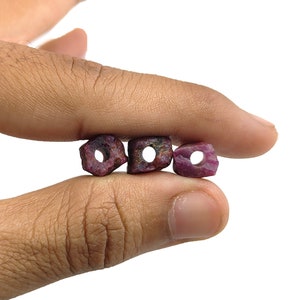 May include: Three rough-cut ruby beads with a hole drilled through each. The beads are held between fingers against a white background. The rubies are a deep, rich red color.