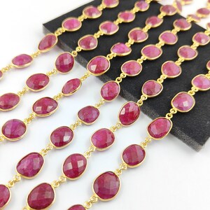 Op de afbeelding: Een streng robijn edelsteen kralen geregen aan een gouden ketting. De kralen zijn ovaalvormig en hebben een facetgeslepen afwerking. De ketting is gemaakt van kleine, ronde schakels.