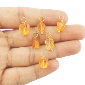 May include: Seven small, raw, orange-colored citrine gemstone pendants with gold-colored jump rings. The pendants are on a hand.