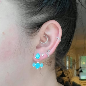 Pode incluir: Um par de brincos de pedra preciosa azul turquesa com forma de lágrima. Os brincos são presos a um poste de prata e têm uma pequena barra de prata que se estende da parte inferior da lágrima. Os brincos são usados no lóbulo da orelha e a barra é colocada atrás da orelha.