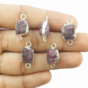 May include: Five raw ruby gemstone pendants with silver-colored metal accents. The rough-cut rubies are a deep reddish-purple color, and each has a silver-colored metal loop at the top and bottom for stringing or attaching to other jewelry components.