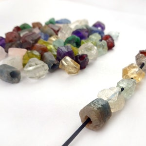 May include: A close-up of a string of colorful gemstone beads. The beads are faceted and have a rough, natural look. The string is made of black thread and is strung with a variety of gemstones, including amethyst, citrine, aquamarine, and garnet.