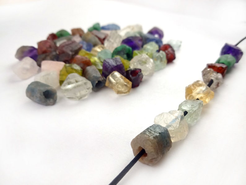 May include: A close-up of a string of colorful gemstone beads. The beads are faceted and have a rough, natural look. The string is made of black thread and is strung with a variety of gemstones, including amethyst, citrine, aquamarine, and garnet.