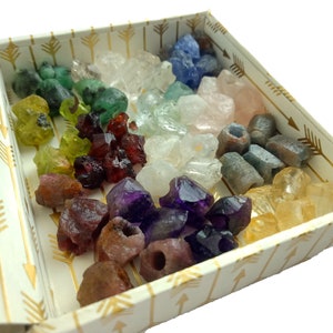 Pu&ograve; includere: Una scatola decorativa piena di una variet&agrave; di pietre preziose grezze colorate. Le pietre preziose includono sfumature di verde, rosso, viola, blu e giallo. La scatola &egrave; bianca con un motivo a frecce dorate.