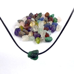 May include: A black cord necklace with a green gemstone pendant. The pendant is a rough, irregular shape. A pile of various colored gemstones is in the background.