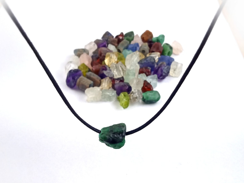 May include: A black cord necklace with a green gemstone pendant. The pendant is a rough, irregular shape. A pile of various colored gemstones is in the background.