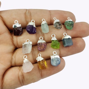 May include: Twelve raw gemstone pendants with silver bail settings. The gemstones are amethyst, aquamarine, ruby, emerald, sapphire, citrine, rose quartz, garnet, peridot, and clear quartz.