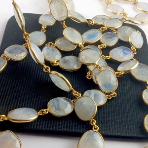 Peut inclure: Un long collier compos&eacute; de perles de pierre de lune ovales avec une cha&icirc;ne dor&eacute;e. Les perles sont de couleur bleu-blanc clair et ont une finition chatoyante.