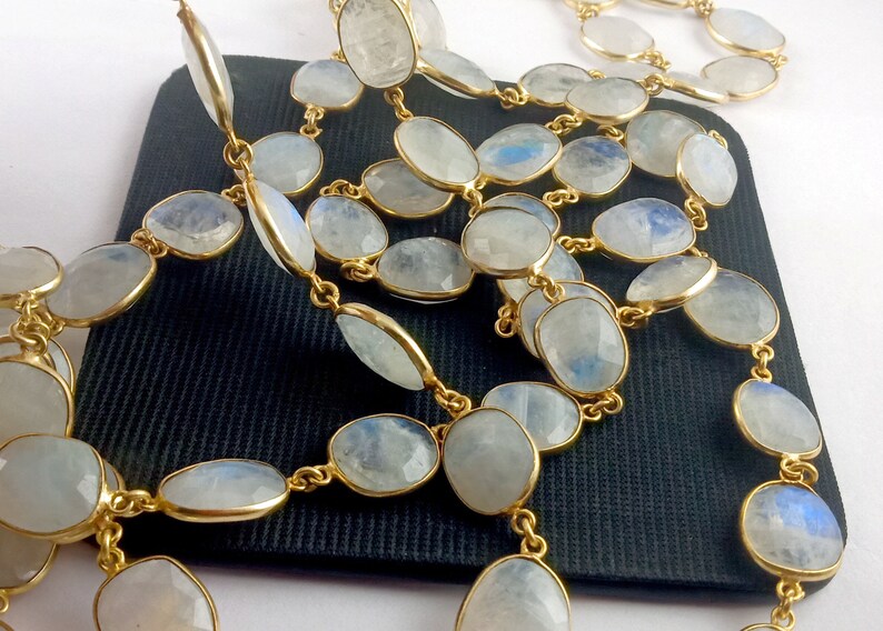 Peut inclure: Un long collier compos&eacute; de perles de pierre de lune ovales avec une cha&icirc;ne dor&eacute;e. Les perles sont de couleur bleu-blanc clair et ont une finition chatoyante.