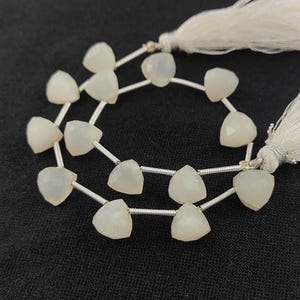 Puede incluir: Una hebra de cuentas de piedra lunar blanca, cada cuenta tiene forma triangular con una superficie facetada. Las cuentas están enhebradas en alambre plateado y tienen un borla en cada extremo.
