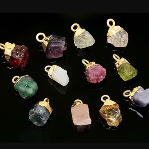 May include: Assortment of 12 gold-plated gemstone pendants. The pendants feature various colors, including red, green, blue, purple, pink, and yellow. Each pendant is shaped like a rough, uncut gemstone.