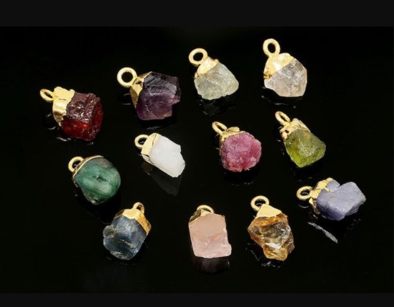 May include: Assortment of 12 gold-plated gemstone pendants. The pendants feature various colors, including red, green, blue, purple, pink, and yellow. Each pendant is shaped like a rough, uncut gemstone.