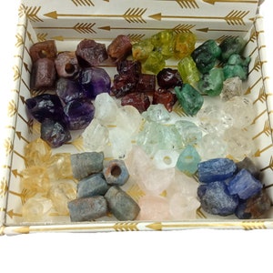 Pu&ograve; includere: Una scatola decorativa piena di una variet&agrave; di pietre preziose grezze nelle tonalit&agrave; di viola, verde, giallo, blu e trasparente. La scatola ha uno sfondo bianco con disegni di frecce dorate.