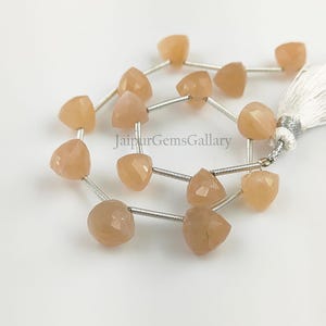 Puede incluir: Una hebra de cuentas de piedras preciosas de color melocotón enhebradas en alambre plateado. Las cuentas son facetadas y tienen forma de corazón. La hebra termina con un borla blanca.