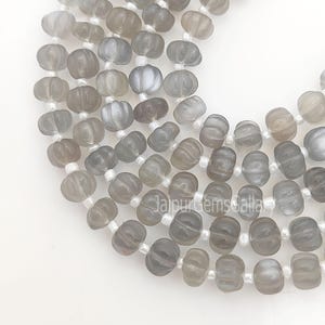 Op de afbeelding: Close-up van strengen grijze edelsteen kralen in de vorm van een pompoen. De kralen zijn geregen op witte draad en hebben een doorschijnend uiterlijk. De kralen hebben grijstinten.