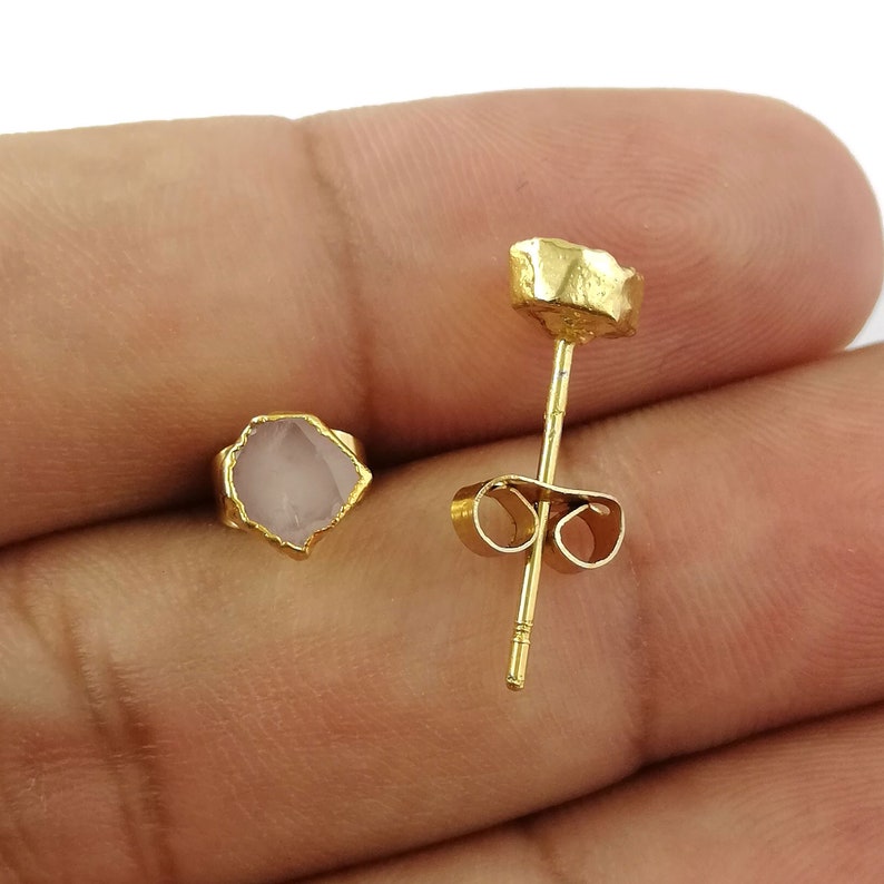May include: A pair of gold earrings with a pink gemstone. The earrings are made of gold and have a simple design. The gemstones are small and have a natural, rough look.