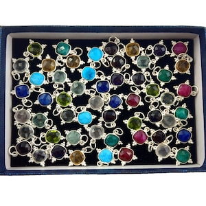 May include: A blue box filled with silver charms featuring various coloured gemstones. The charms are arranged in a random pattern and include colours such as turquoise, green, red, purple, and clear.