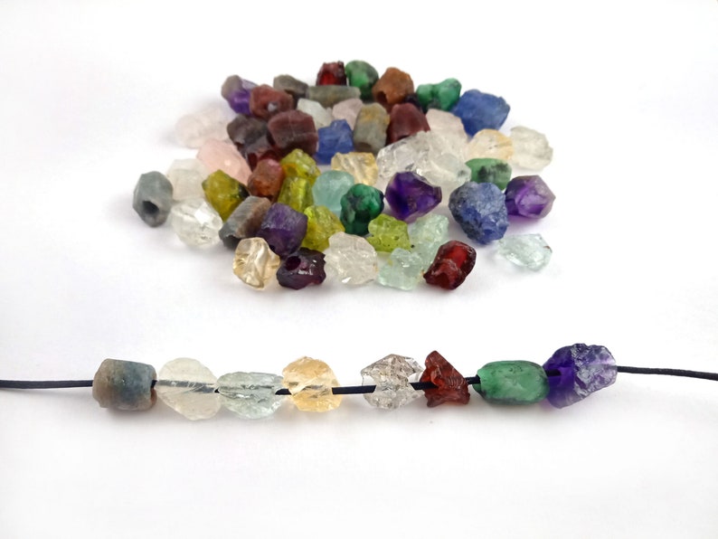 May include: A variety of colorful gemstone beads, including amethyst, aquamarine, citrine, emerald, garnet, and ruby, are scattered on a white surface. A strand of beads is strung on a black cord, showcasing the different colors and textures of the gemstones.