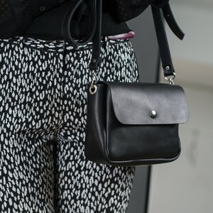 Może przedstawiać: Czarna skórzana torebka na ramię z klapką i srebrnym guzikiem. Torebka jest noszona na czarno-białej wzorzystej koszuli.