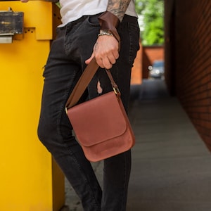 Puede incluir: Una persona con jeans negros y una camiseta blanca sostiene un bolso de mensajero de cuero marrón con una hebilla dorada. El bolso se lleva sobre el hombro de la persona. La persona está de pie en una acera.