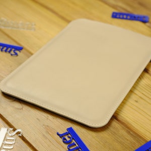 May include: A beige leather tablet sleeve with a stitched edge. The sleeve is lying on a wooden surface.