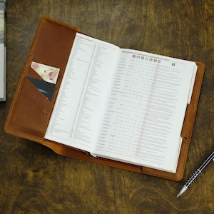 May include: A tan leather notebook cover featuring internal pockets holding two small cards. The cover is open, displaying a white paper planner with printed text and a table of data. A black pen rests on the surface next to the leather notebook.