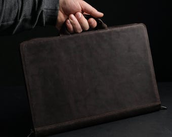 May include: A dark brown leather portfolio case with a smooth, matte finish and a top handle. The rectangular document holder features a zippered closure along the edges, providing a secure and structured design for carrying papers or a slim laptop.