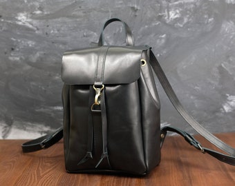 Può includere: Uno zaino in pelle nera con patta ripiegabile, chiusura con gancio in metallo color ottone, manico superiore e spallacci regolabili. La borsa presenta una finitura liscia, una forma rettangolare strutturata e dettagli con cinturini verticali.