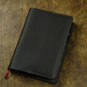 May include: A black leather journal with a red ribbon bookmark. The journal is closed and lies on a wooden surface. The cover is smooth and the edges are neatly stitched.