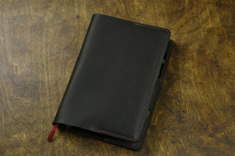 May include: A black leather journal with a red ribbon bookmark. The journal is closed and lies on a wooden surface. The cover is smooth and the edges are neatly stitched.