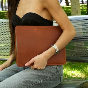 May include: A rectangular, smooth, tan leather laptop sleeve with rounded corners. The sleeve is held horizontally, showcasing a clean, minimalist design with visible stitching along the edges and a natural, matte finish on the leather surface.