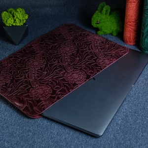 K&ouml;nnte beinhalten: Eine dunkelrote Lederh&uuml;lle f&uuml;r Laptops mit einem gepr&auml;gten Blumenmuster. Die H&uuml;lle ist geschlossen und ein silberner Laptop befindet sich im Inneren.