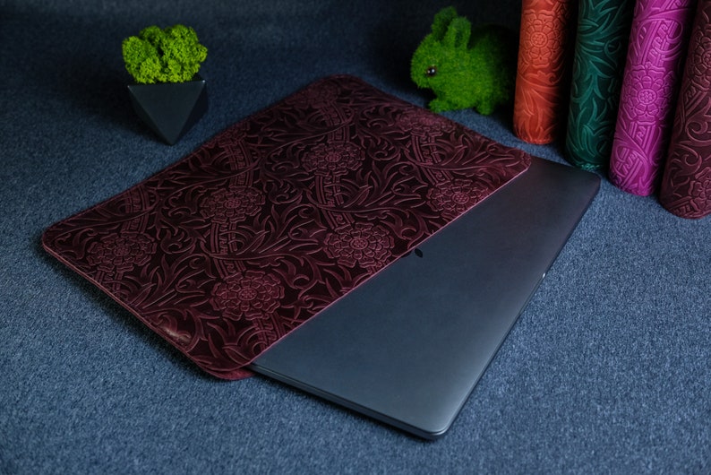 K&ouml;nnte beinhalten: Eine dunkelrote Lederh&uuml;lle f&uuml;r Laptops mit einem gepr&auml;gten Blumenmuster. Die H&uuml;lle ist geschlossen und ein silberner Laptop befindet sich im Inneren.