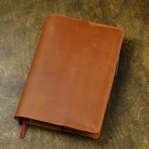 May include: A tan leather notebook cover with a smooth, matte finish and visible stitching along the edges. The cover features a single vertical fold and a small, thin red ribbon bookmark extending from the bottom edge of the closed notebook.