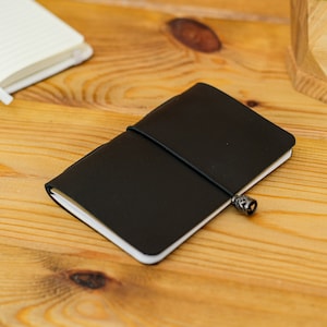 May include: A black leather journal with a strap closure and decorative metal charm. The notebook is closed and lies on a wooden surface, with a pen and open notebook visible in the background.