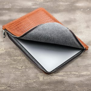 Könnte beinhalten: Eine hellbraune Laptoptasche mit einer strukturierten Krokodilleder-Optik auf der Außenseite und einem weichen grauen Filzfutter. Die Tasche verfügt über einen silbernen Metallreißverschluss und enthält einen flachen, silbernen Laptop.