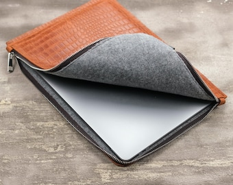 May include: A tan laptop sleeve with a textured crocodile skin pattern on the exterior and a soft gray felt lining. The sleeve features a silver metal zipper closure and contains a thin, metallic silver laptop inside the main compartment.