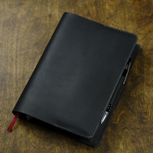 May include: A black leather journal with a pen tucked into the spine, resting on a wooden surface. The journal has a red ribbon bookmark. The pen is silver and black.