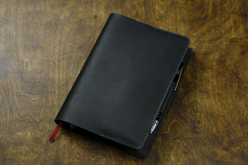 May include: A black leather journal with a pen tucked into the spine, resting on a wooden surface. The journal has a red ribbon bookmark. The pen is silver and black.