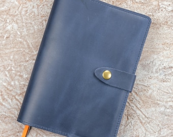 May include: A navy blue leather notebook cover featuring a smooth matte finish, a single strap closure secured with a round brass-coloured metal snap button, and a small orange ribbon bookmark extending from the bottom edge.
