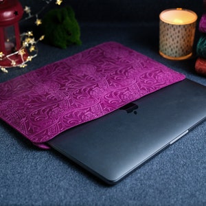 K&ouml;nnte beinhalten: Eine violette Laptoptasche aus Leder mit einem gepr&auml;gten Blumenmuster. Die Tasche ist geschlossen und ein Laptop befindet sich im Inneren.