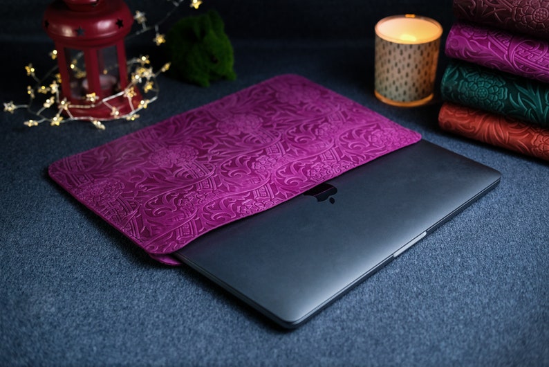 K&ouml;nnte beinhalten: Eine violette Laptoptasche aus Leder mit einem gepr&auml;gten Blumenmuster. Die Tasche ist geschlossen und ein Laptop befindet sich im Inneren.