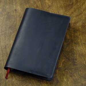 May include: A dark navy blue leather notebook cover with a smooth, matte finish and clean stitched edges. A small, thin red ribbon bookmark extends from the bottom left corner of the rectangular cover, providing a subtle contrast to the dark surface.