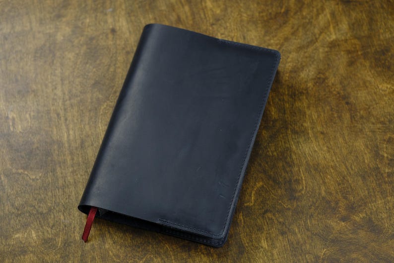 May include: A dark navy blue leather notebook cover with a smooth, matte finish and clean stitched edges. A small, thin red ribbon bookmark extends from the bottom left corner of the rectangular cover, providing a subtle contrast to the dark surface.