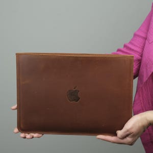 Może przedstawiać: Prostokątne etui na laptopa z brązowej skóry o gładkim, matowym wykończeniu z widocznymi szwami wzdłuż krawędzi. Na środku etui znajduje się wytłoczone logo Apple, które stanowi subtelny kontrast dla powierzchni z brązowej skóry.